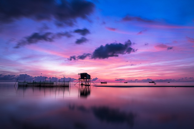 Stunning pink and blue sunset over calm sea, with a silhouette of a fishing hut on stilts reflecting on the water.