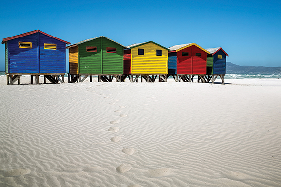 Colorful beach huts on a sandy shore under a clear blue sky, with ocean waves in the background.