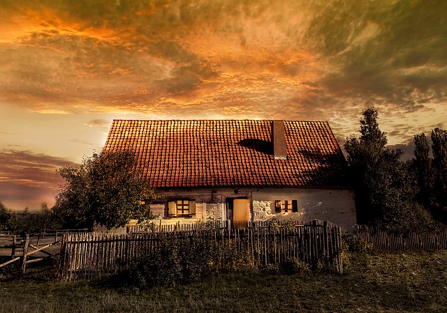 Rustic cottage with red roof under dramatic sunset sky, surrounded by wooden fence and trees.