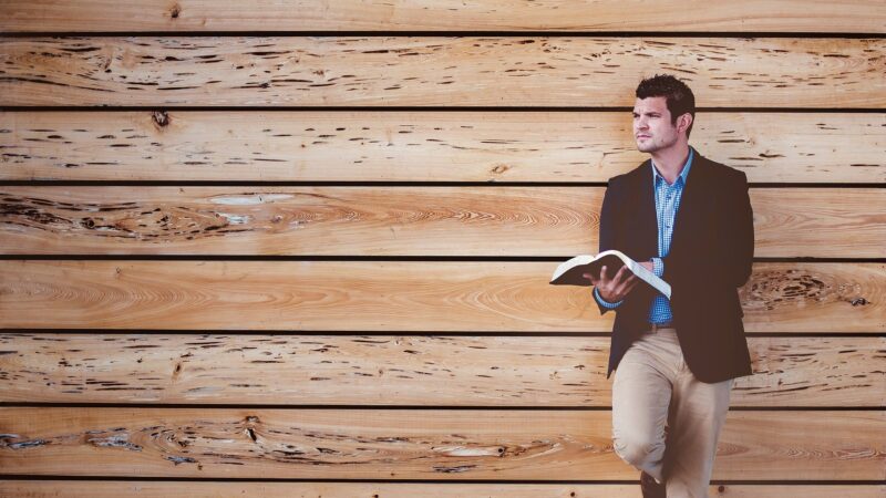 Man in casual attire reading against wood wall.