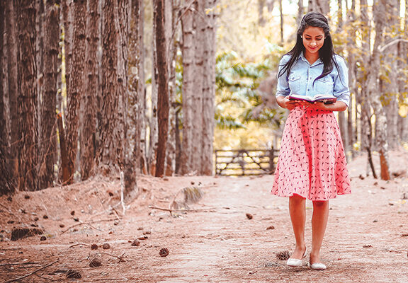 Woman reading in a forest, wearing a denim jacket and pink skirt, surrounded by tall trees and autumn leaves.