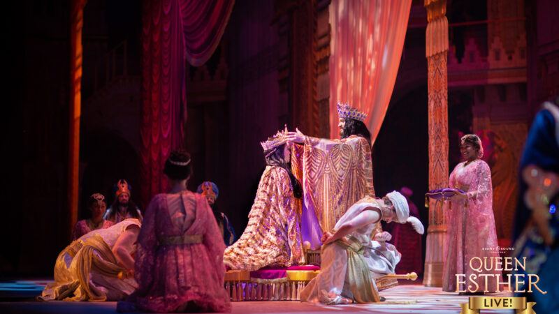 Theatrical scene from Queen Esther featuring actors in royal costumes on stage, with elaborate set design.