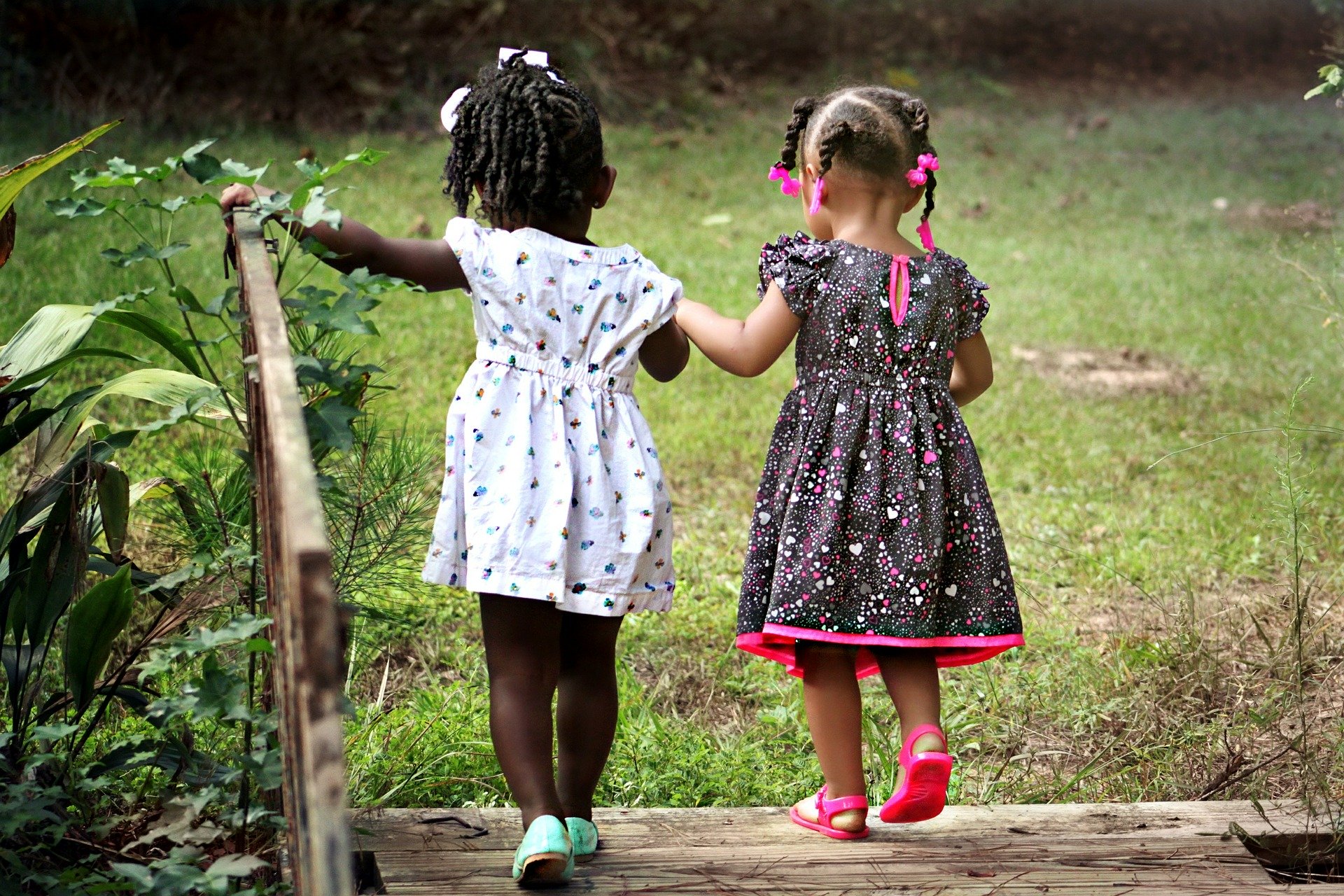 Two young girls holding hands and walking on a wooden path in a grassy park, dressed in summer dresses.
