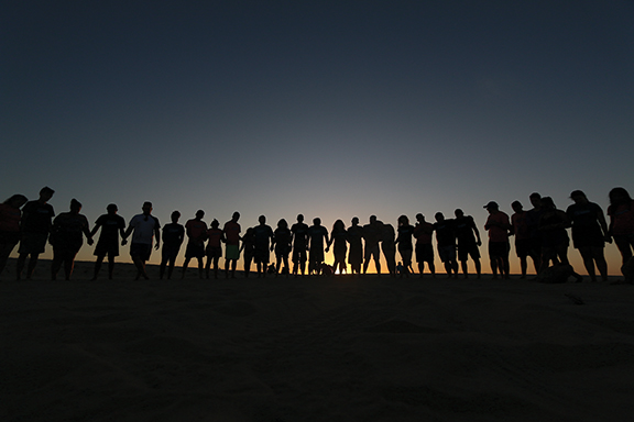 Silhouettes of people holding hands in a circle at sunset, symbolizing unity and togetherness on a beach.