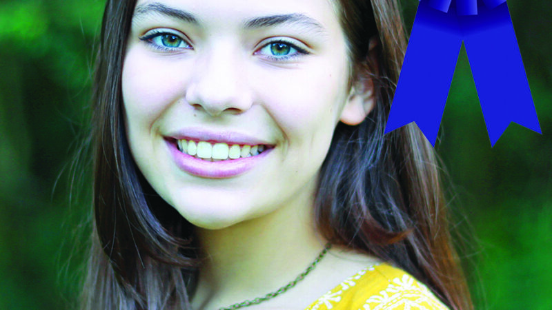 Smiling woman with long hair, wearing a yellow top, a blue First Place ribbon beside her, green background.