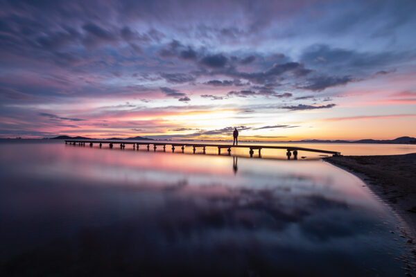 Serene sunset over a calm lake with a silhouette standing on a long pier.