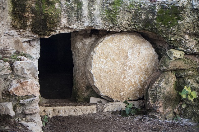 Ancient stone tomb with round entrance, partially covered by a large stone, surrounded by rocks and vegetation.