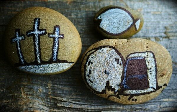 Painted stones with Christian symbols: three crosses, an angel, and a fish on a wooden surface.