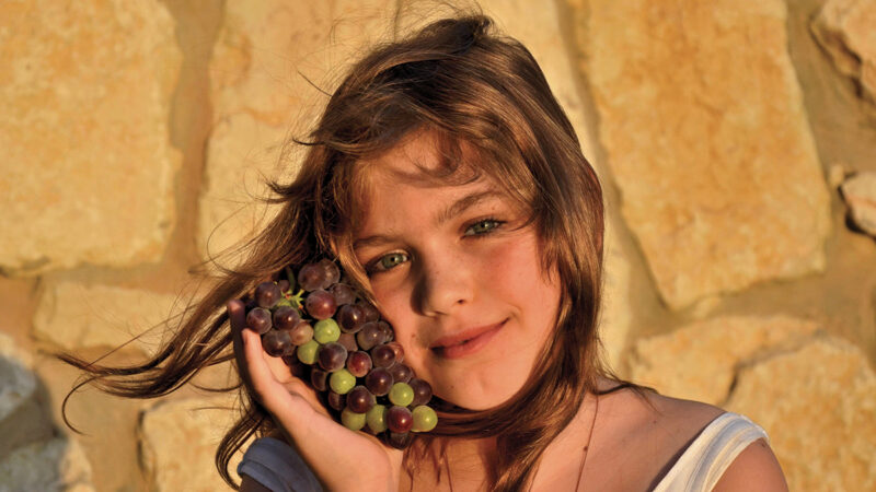 Girl smiling with grapes against stone wall in warm sunlight.
