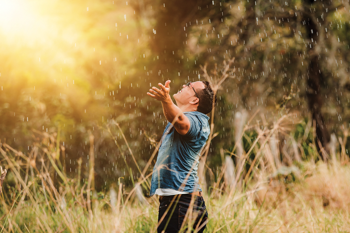 Man joyfully embracing rain in sunlit field, arms outstretched, surrounded by grass and trees.