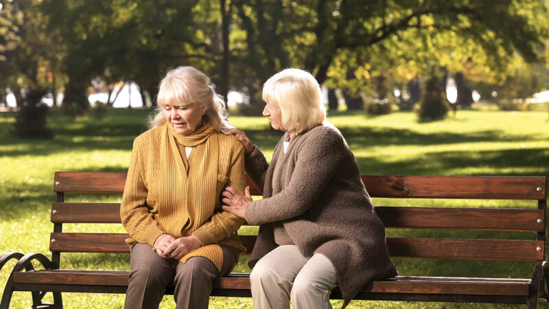 Two elderly women support each other on a park bench, surrounded by lush greenery and sunlight.