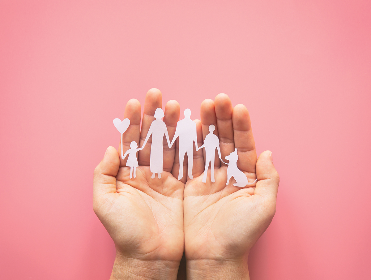 Hands holding paper cut-out family and dog on a pink background, symbolizing love and protection.