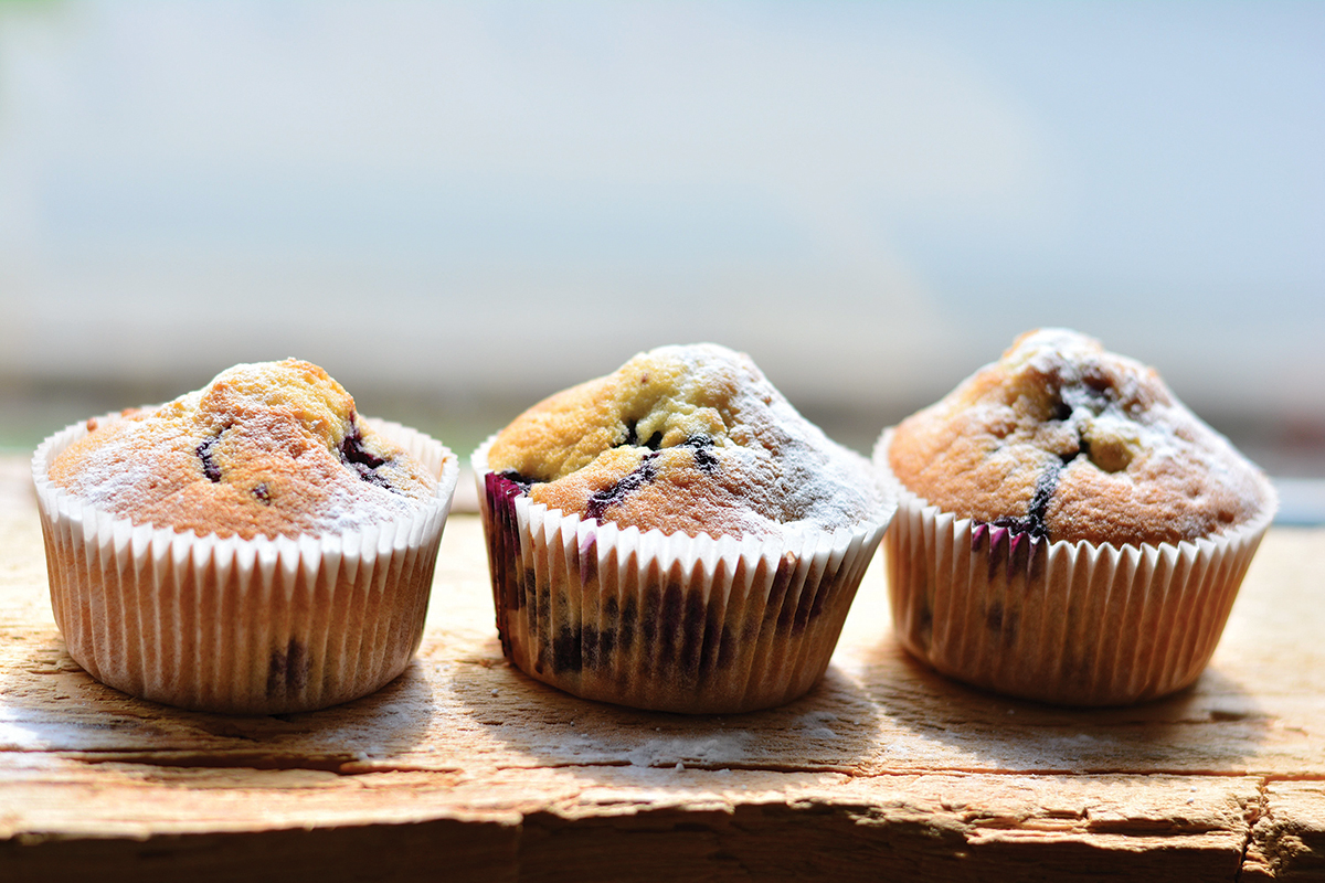 Three freshly baked blueberry muffins dusted with powdered sugar on a wooden surface.