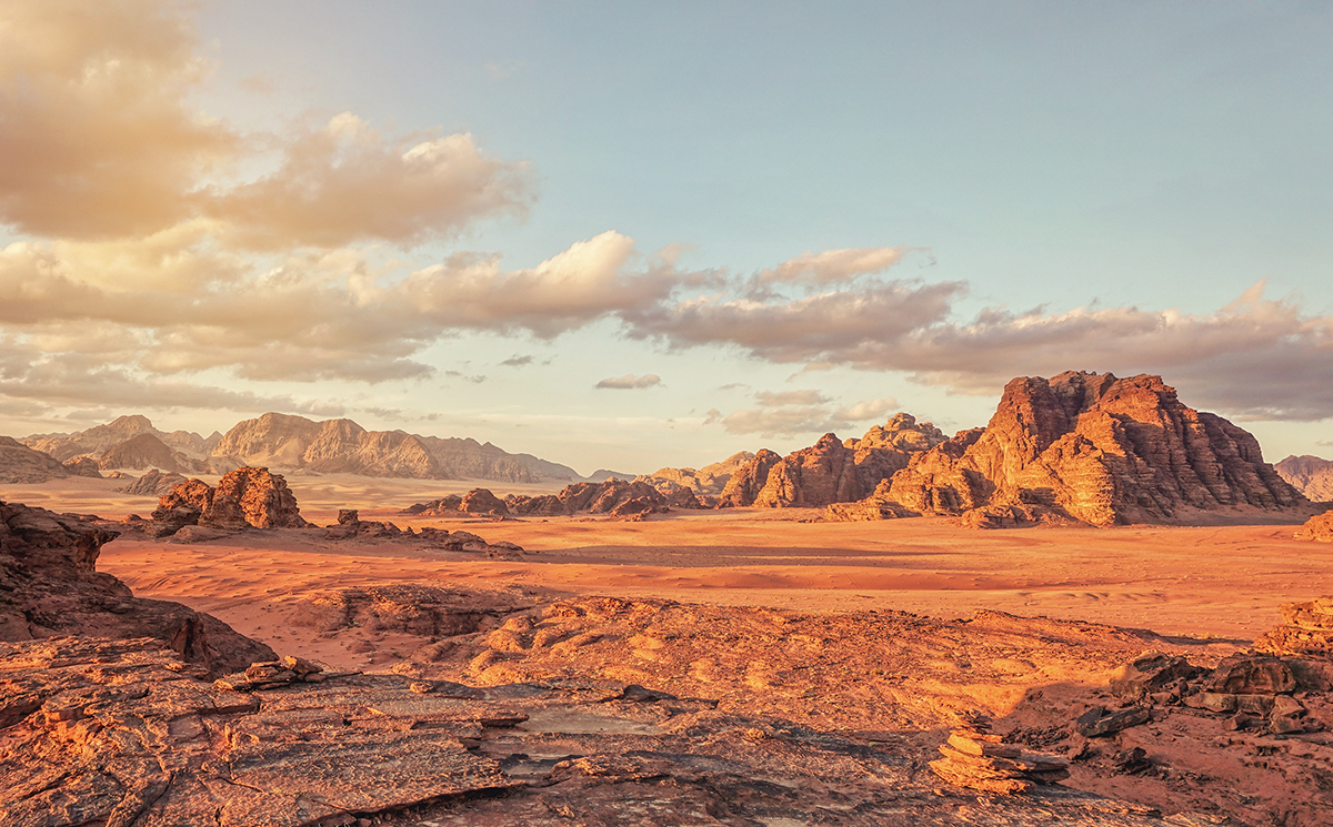 Stunning desert landscape with rocky mountains, golden sand, and cloudy sky at sunset.