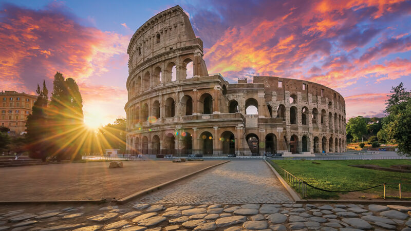 Sunset view of the Colosseum in Rome, Italy, with vibrant sky and sunlight peeking through the ruins.