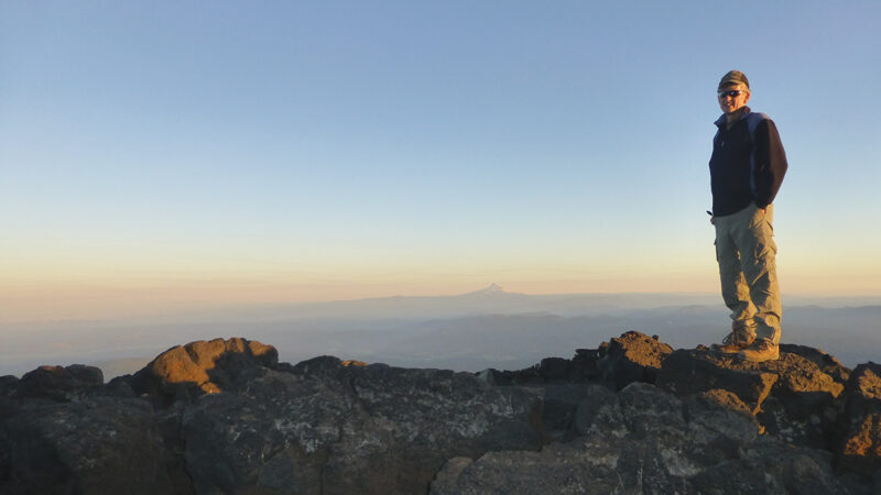 Person standing on rocky mountain summit during sunrise, overlooking distant mountain landscape.