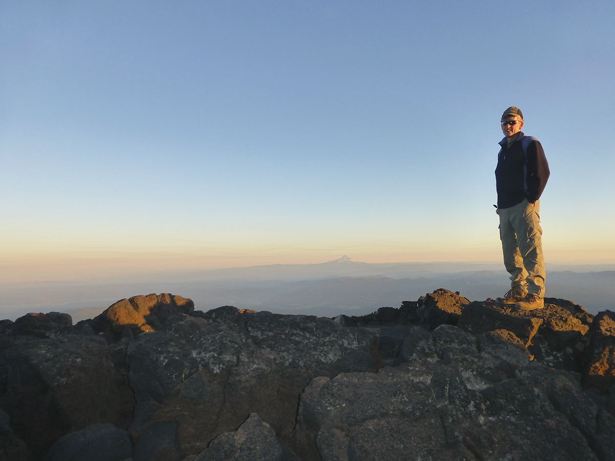 Person standing on rocky mountain summit during sunrise, overlooking distant mountain landscape.