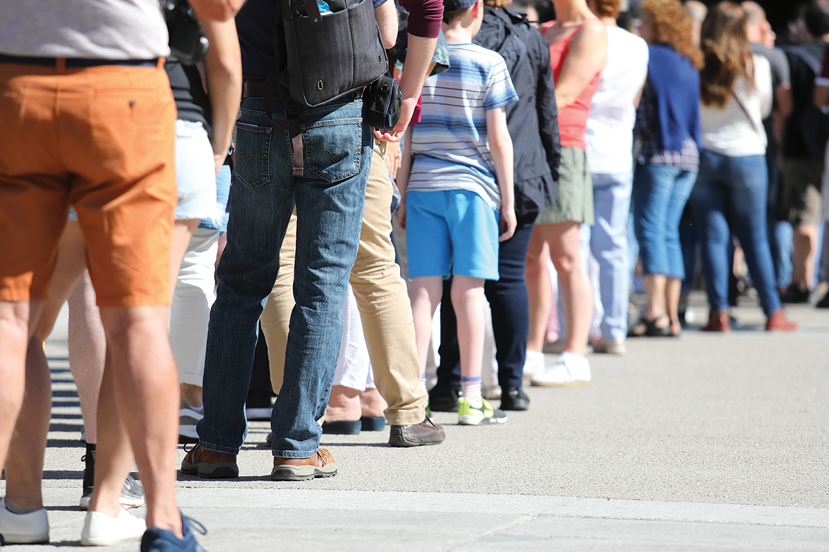 A diverse group of people standing in line outdoors on a sunny day, showing casual summer clothing and footwear.