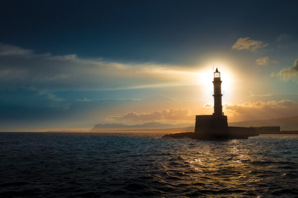Lighthouse at sunset over ocean waves, casting a silhouette against dramatic clouds and distant mountains.