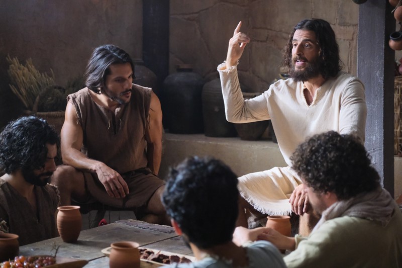 Biblical scene with a man teaching a group around a table in ancient attire, discussing with gestures and focus.