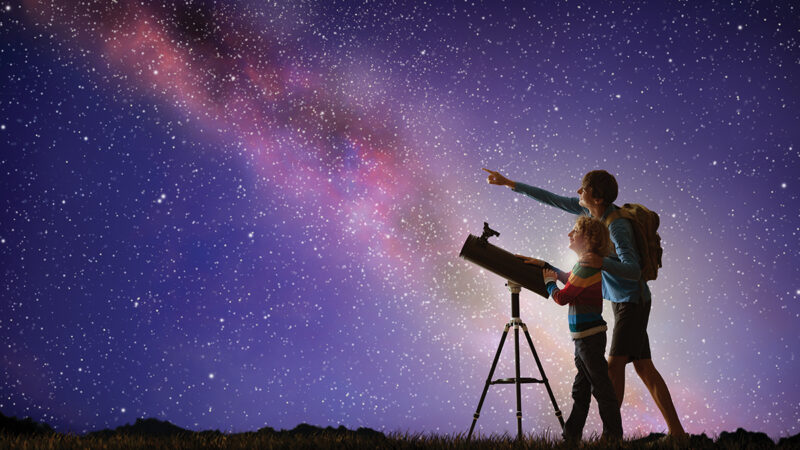 Father and child stargazing with telescope under a starry sky.