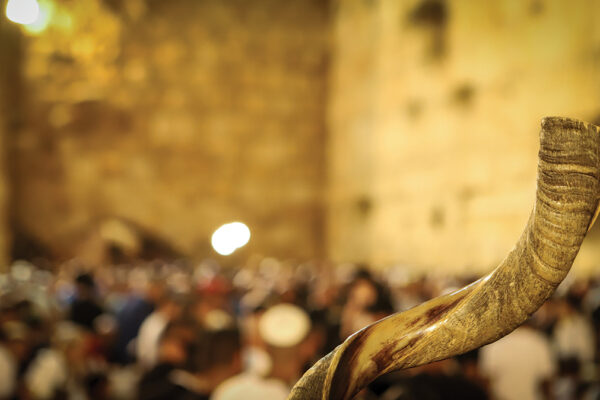 Shofar horn in focus against a blurred background of a stone wall and crowd at a religious gathering.