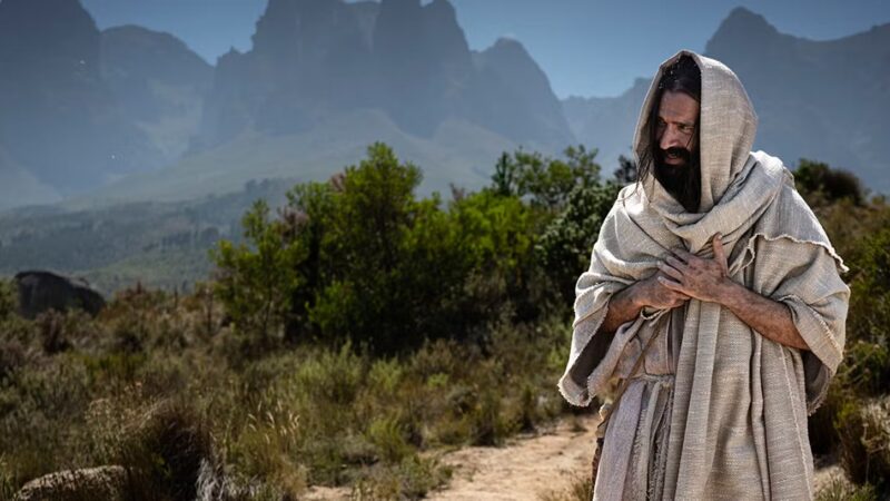 Man standing in desert landscape with mountains in the background, wearing traditional robe and hood.