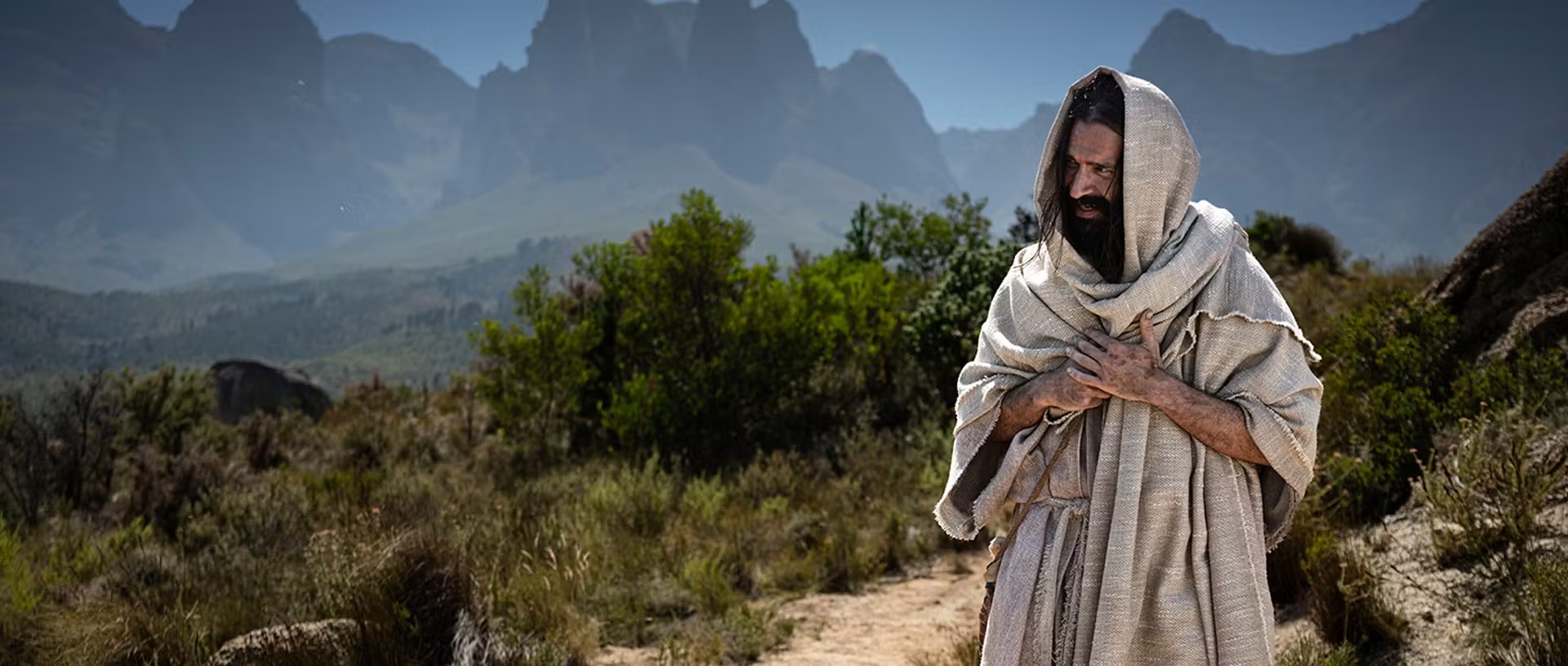 Man standing in desert landscape with mountains in the background, wearing traditional robe and hood.