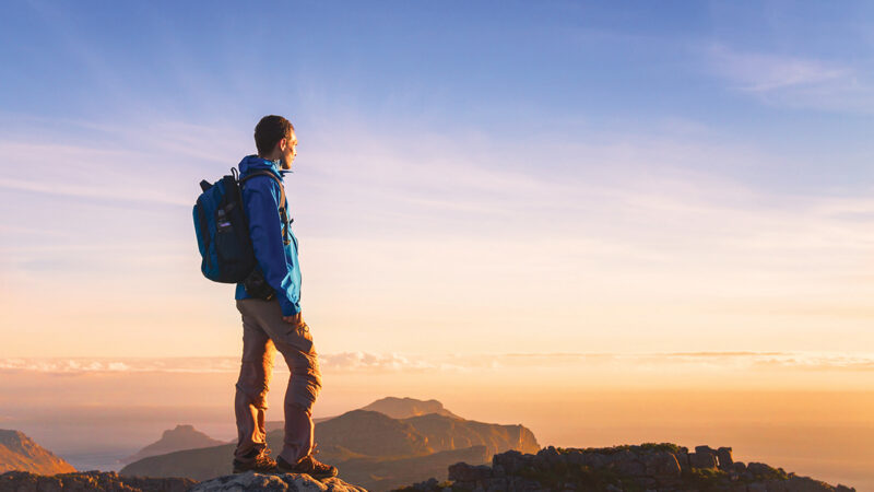 Hiker with backpack stands on rocky cliff, gazing at sunrise over mountains and sea, under a clear blue sky.