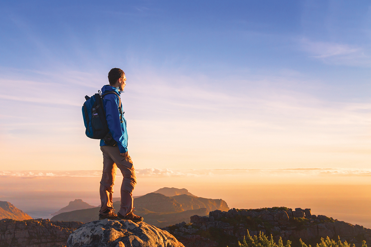 Hiker with backpack stands on rocky cliff, gazing at sunrise over mountains and sea, under a clear blue sky.