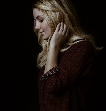 A woman with long blonde hair poses against a dark background, touching her ear while wearing a diamond ring.