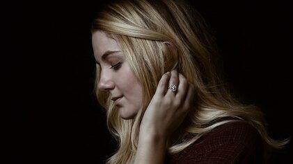 A woman with long blonde hair poses against a dark background, touching her ear while wearing a diamond ring.