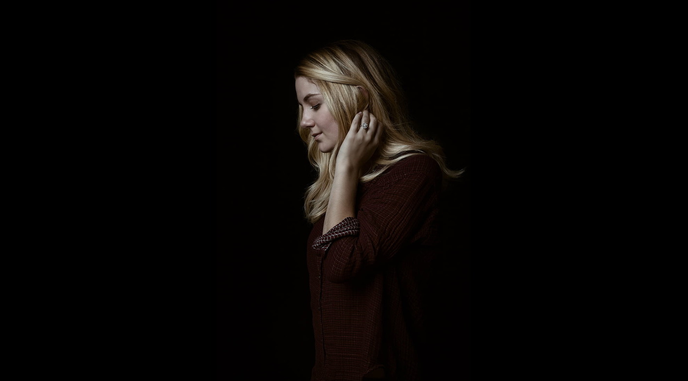 Alt text: Woman with blonde hair in side profile against dark background, wearing a dark shirt, hand touching her hair.