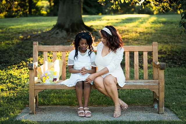Mother and daughter in white dresses sitting on a park bench with flowers, surrounded by greenery and soft sunlight.