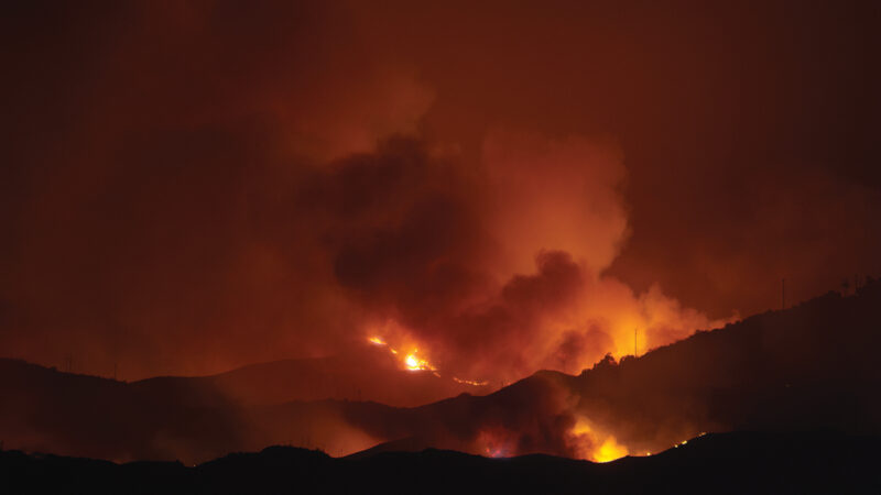Wildfire raging at night, illuminating dark clouds and landscape with intense orange flames and thick smoke.