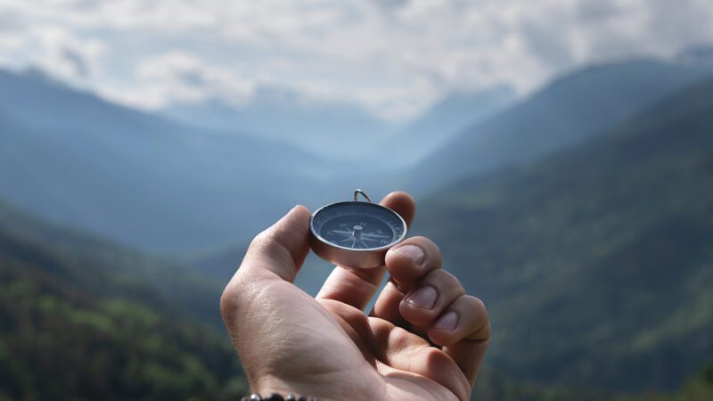 Magnetic compass in the palm of a male hand against the backdrop of a mountain range in the clouds in the summer outdoors, travel, first person view